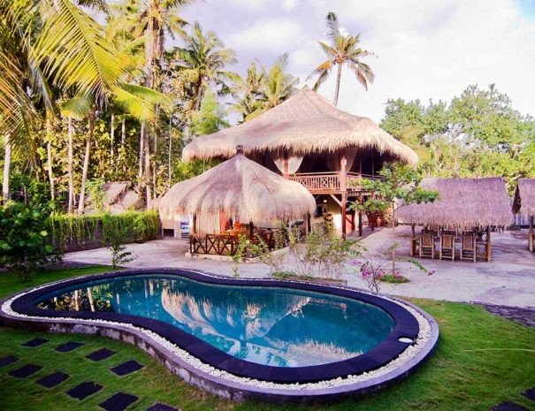 Picture of the outside of a diving resort, with the main building, outside tables and a swimming pool. 