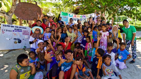 Picture of a big group of adults and children outside a dive centre. They are holding banners and waving at the camera. 