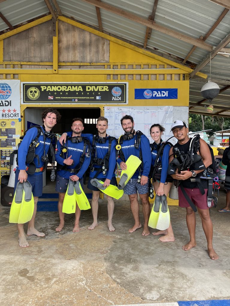 5 divers and a guide posing with yellow fins in front of Panorama Diver shop sign